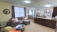 This single-story residence showcases an open-concept floor plan that integrates the living, dining, and kitchen areas into one cohesive, flowing space. The living area is anchored by plush carpeting and features large windows dressed with gray curtains and horizontal blinds, allowing generous natural light to fill the room. A comfortable sectional sofa with colorful accent pillows faces a stylish round glass coffee table, creating an inviting focal point. Transitioning toward the kitchen, a dark wood breakfast bar with light stone countertops serves as a natural divider, complemented by a modern pendant light overhead. The kitchen island provides additional functionality and visual contrast against the lighter walls and flooring. Beyond the living area, the dining space benefits from a bay window configuration, further enhancing the sense of openness and light throughout. A vibrant piece of wall art adds a pop of color and personality to the space. The overall ambiance is modern and welcoming, combining warm tones, thoughtful lighting, and an efficient layout suited for both everyday living and entertaining.