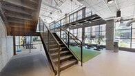 This two-level fitness facility showcases a striking industrial aesthetic defined by exposed concrete ceilings, raw concrete columns, concrete masonry unit walls, and visible ductwork and mechanical systems. Large, grid-paned windows line the street-facing facade on both levels, flooding the interior with abundant natural light and offering open views of the surrounding urban neighborhood.

The lower level serves as the primary fitness floor, offering generous open space divided into distinct workout zones. Cardio equipment, including a row of treadmills, is positioned directly in front of the expansive windows, while dedicated strength training areas feature free weights, benches, rowing machines, and cable machines. Rubber and turf flooring define different training zones, providing both functionality and visual variety. A steel staircase with industrial mesh railings connects the two levels.

The upper level features a light-filled mezzanine walkway with natural hardwood flooring running along the perimeter, framed by the same mesh-panel railings and offering overlook views of the fitness floor below. A separate studio space on this level features wide-plank hardwood floors and mirrored walls, providing an ideal setting for group fitness classes or yoga. Large overhead fans and pendant lighting complement the industrial character throughout. The overall design balances raw, utilitarian materials with thoughtful functionality.