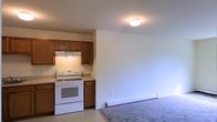 This single-floor space offers a practical and straightforward layout with an open-concept design that connects the kitchen and living area. The kitchen is equipped with warm-toned wood cabinetry providing ample storage, a white electric range with an overhead ventilation hood, and light-colored countertops. The kitchen flooring transitions seamlessly into the carpeted living area, which offers a comfortable and functional space. A baseboard heating unit runs along the living area wall, ensuring efficient climate control. Flush-mounted ceiling lighting illuminates both areas. The neutral wall tones throughout create a clean, versatile backdrop suitable for a variety of furnishing styles. The open flow between the kitchen and living space makes the overall layout feel cohesive and easy to navigate.