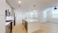 This single-floor residence showcases a cohesive modern design highlighted by light hardwood flooring that runs throughout the entire space, crisp white walls, and abundant natural light from multiple windows dressed with clean horizontal blinds. The open-concept kitchen serves as the centerpiece, anchored by a generous white quartz island with an integrated sink and matte black fixtures. Pendant lights hang above the island, complementing recessed lighting overhead. Light wood cabinetry lines the walls alongside a subway tile backsplash, while stainless steel appliances — including a refrigerator with ice maker, gas range, and built-in microwave — provide a polished, functional finish. A built-in beverage cooler adds an upscale touch.

The bedrooms are bright and well-proportioned, each featuring ceiling fans with tray ceiling details and multiple windows that invite natural light. One bedroom includes a walk-in closet with built-in shelving, leading directly into a private en-suite bathroom. The bathrooms are elegantly appointed with large-format tile flooring, dark-toned vanities with white countertops, matte black hardware, and a floor-to-ceiling marble-look tiled walk-in shower with a glass enclosure. Overall, the space presents a clean, contemporary aesthetic with quality finishes throughout.