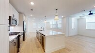 This single-floor residence showcases a cohesive modern design highlighted by light hardwood flooring that runs throughout the entire space, crisp white walls, and abundant natural light from multiple windows dressed with clean horizontal blinds. The open-concept kitchen serves as the centerpiece, anchored by a generous white quartz island with an integrated sink and matte black fixtures. Pendant lights hang above the island, complementing recessed lighting overhead. Light wood cabinetry lines the walls alongside a subway tile backsplash, while stainless steel appliances — including a refrigerator with ice maker, gas range, and built-in microwave — provide a polished, functional finish. A built-in beverage cooler adds an upscale touch.

The bedrooms are bright and well-proportioned, each featuring ceiling fans with tray ceiling details and multiple windows that invite natural light. One bedroom includes a walk-in closet with built-in shelving, leading directly into a private en-suite bathroom. The bathrooms are elegantly appointed with large-format tile flooring, dark-toned vanities with white countertops, matte black hardware, and a floor-to-ceiling marble-look tiled walk-in shower with a glass enclosure. Overall, the space presents a clean, contemporary aesthetic with quality finishes throughout.