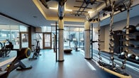 Located at 600 S Wells St in Chicago's vibrant South Loop neighborhood, this professionally equipped fitness center offers a comprehensive workout environment across a single well-designed floor. The space is flooded with natural light through expansive floor-to-ceiling glass walls, framing views of the surrounding Chicago cityscape and an adjacent outdoor terrace area.

The cardio zone is outfitted with a curved manual treadmill, Peloton stationary bikes, and rowing machines, all positioned to take full advantage of the panoramic city views. Overhead, an industrial-style functional training rig with cables, suspension trainers, and pull-up bars anchors the central training area, supported by structural columns that define each zone.

A dedicated free weights section features a full rack of dumbbells alongside a flat bench, set against a richly textured dark wood accent wall that adds a warm, sophisticated contrast to the otherwise sleek, modern aesthetic. Full-length mirrors expand the visual depth of the space and support form-focused training.

Toward the rear, a separate room is designated for combat and high-intensity training, complete with a heavy punching bag, plyo boxes, and ample open floor space with dark rubberized flooring. Recessed and linear LED lighting throughout maintains a clean, modern atmosphere well-suited for focused, high-performance training.