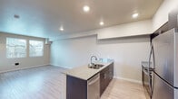 This single-floor unit offers a clean, contemporary design with an open-concept layout that seamlessly connects the kitchen and living area. The kitchen serves as the centerpiece, featuring a peninsula with a double stainless steel sink and granite countertops, dark espresso cabinetry with brushed hardware, and a full suite of stainless steel appliances including a refrigerator, electric range, and over-the-range microwave. Light wood-style flooring runs throughout the main living area, enhancing the sense of openness and warmth. Recessed lighting illuminates the space efficiently, complementing the neutral, light-toned walls. The spacious living area benefits from a window that invites natural light and offers views of the surrounding neighborhood. The bedroom is generously sized with plush carpeting and a bank of large windows fitted with blinds, providing abundant natural light and outdoor views. The bathroom is well-appointed with a granite-topped dark vanity, a large mirror, a combination tub and shower, and light wood-style flooring that mirrors the rest of the unit. Overall, the space presents a cohesive, modern aesthetic with functional finishes suited for comfortable everyday living.