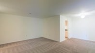 This single-floor space features a spacious, open main living area characterized by neutral-toned wall-to-wall carpeting and crisp white walls that create a bright, clean atmosphere. The room offers generous square footage, providing ample flexibility for a variety of furniture arrangements and layouts. The standard ceiling height contributes to a comfortable, functional feel throughout the space. A partial wall divides the main living area from an adjoining kitchen, which is visible through an open doorway. The kitchen features warm wood-toned cabinetry and light countertops, offering a practical and functional cooking space. The transition between the carpeted living area and the hard flooring of the kitchen is clearly defined by this architectural division. Electrical outlets are conveniently distributed along the baseboards of the main room, and a ceiling vent is present, indicating a central air system. The overall palette of the space — neutral carpeting, white walls, and natural wood cabinetry — creates a cohesive, understated aesthetic that serves as a versatile canvas for personalization. The layout is straightforward and efficient, making it well-suited for everyday living.