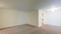 This single-floor space features a spacious, open main living area characterized by neutral-toned wall-to-wall carpeting and crisp white walls that create a bright, clean atmosphere. The room offers generous square footage, providing ample flexibility for a variety of furniture arrangements and layouts. The standard ceiling height contributes to a comfortable, functional feel throughout the space. A partial wall divides the main living area from an adjoining kitchen, which is visible through an open doorway. The kitchen features warm wood-toned cabinetry and light countertops, offering a practical and functional cooking space. The transition between the carpeted living area and the hard flooring of the kitchen is clearly defined by this architectural division. Electrical outlets are conveniently distributed along the baseboards of the main room, and a ceiling vent is present, indicating a central air system. The overall palette of the space — neutral carpeting, white walls, and natural wood cabinetry — creates a cohesive, understated aesthetic that serves as a versatile canvas for personalization. The layout is straightforward and efficient, making it well-suited for everyday living.
