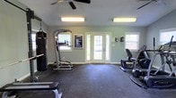Located at 2038 Quail Forest Dr in Raleigh, NC, this fitness center offers a well-equipped, single-floor workout space designed to support a variety of exercise routines. The room features rubber flooring throughout, providing a durable and slip-resistant surface ideal for both cardio and strength training activities. Natural light fills the space through multiple windows and a set of glass-paneled double doors, creating a bright and welcoming atmosphere. Overhead fluorescent lighting supplements the natural light, ensuring the space is well-lit at all times. A ceiling fan adds comfort by promoting air circulation during workouts. The facility is equipped with a range of exercise equipment, including a heavy punching bag, a multi-function power tower for bodyweight exercises, a stationary recumbent bike, and an elliptical machine, along with free weights and a weight bench. The walls are painted in a calming sage green, contributing to a clean and focused environment. Community signage is posted near the entrance, reflecting the shared-use nature of the space. Overall, this fitness center provides a practical and comfortable setting for residents to maintain an active lifestyle.