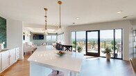 This expansive single-floor communal space is thoughtfully designed to offer a variety of functional zones within an open, light-filled layout. Warm hardwood flooring flows continuously throughout, unifying each area while large windows and glass doors frame sweeping panoramic views of the surrounding landscape, filling the interior with abundant natural light.

The dining and gathering area features a substantial white island beneath brass-accented pendant lights, complemented by a tray ceiling with additional lighting nearby. Multiple marble-topped café tables paired with cushioned seating offer flexible options for casual dining or socializing. A bold, large-scale abstract metal wall art installation serves as a striking focal point, adding an artistic dimension to the space.

The kitchenette is equipped with a stainless steel French-door refrigerator, white shaker cabinetry with gold hardware, quartz countertops, and a vibrant teal zellige tile backsplash that introduces a lively pop of color.

A dedicated game room features a coffered ceiling with a statement chandelier, wall sconces, a mounted flat-screen television, and a full-length shuffleboard table. The lounge area offers plush sectional seating and teal accent chairs positioned directly before floor-to-ceiling windows, maximizing the scenic treetop and hillside views. An exterior terrace, accessible through glass doors, extends the living space outdoors.