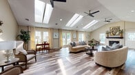 This thoughtfully designed single-floor space offers an open, airy layout highlighted by vaulted ceilings and an impressive array of skylights that flood the interior with abundant natural light. Wide-plank hardwood flooring in warm, multi-toned hues runs continuously throughout, unifying the various living areas with a cohesive, polished look.

The expansive main living area is anchored by a fireplace and comfortably accommodates multiple seating arrangements, enhanced by ceiling fans and recessed lighting. Large windows and glass-paneled doors line the perimeter, offering generous views of the surrounding greenery and allowing natural light to pour in from multiple directions.

The dining area, situated adjacent to the living space, features vaulted ceilings and corner windows that create a bright, welcoming setting. The connected kitchen is well-appointed with warm honey-toned cabinetry, light granite countertops, and stainless steel appliances, providing ample storage and workspace.

Throughout the space, neutral wall tones complement the natural wood flooring, creating a calm and versatile backdrop. The open floor plan promotes a seamless flow between the living, dining, and kitchen zones, making the space both functional and inviting. Overall, the combination of skylights, vaulted ceilings, and generous fenestration gives this space a notably bright and expansive character.