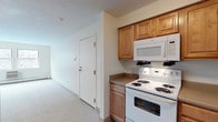 This single-floor residence offers three bedrooms and two full bathrooms, designed with a practical and comfortable layout. The kitchen is well-appointed with warm-toned wooden cabinetry providing generous storage, a full suite of white appliances including an electric range with over-the-range microwave, dishwasher, and refrigerator, complemented by neutral countertops and easy-care tile flooring. The living area is open and airy, featuring soft neutral carpet, baseboard heating, and a windowed door that invites natural light. Multiple windows throughout the bedrooms allow for ample daylight and views of the surrounding exterior, while the carpeted floors lend a cozy, comfortable feel to each room. The two bathrooms are clean and functional — one features a tub/shower combination with white tile surround, a vanity with integrated sink, and stone-look tile flooring; the second offers a standalone glass-door shower stall, a white vanity with mirrored medicine cabinet, and matching tile flooring. The overall aesthetic is neutral and move-in ready, with a straightforward floor plan that connects all rooms efficiently for easy everyday living.