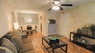 This single-floor residence offers an open and connected layout that seamlessly integrates the living, dining, and kitchen areas into one cohesive space. The living room is anchored by a ceiling fan with integrated lighting, providing both comfort and illumination. Hardwood-style flooring runs throughout the main living areas, adding warmth and a clean, consistent aesthetic. A glass-topped coffee table and comfortable sectional seating define the living space, while a dedicated dining area features a round table and chairs beneath a pendant light fixture, creating an inviting spot for meals. The kitchen is accessible via a raised breakfast bar or pass-through counter, which houses a microwave and provides a functional divide between cooking and living spaces. The kitchen itself is partially visible, suggesting additional cabinetry and counter space beyond. Neutral beige and sage green tones on the walls contribute to a calm and welcoming ambiance throughout. A white paneled entry door adds a classic touch to the interior. The open layout allows natural sightlines across the entire main living zone, making the space feel airy and well-connected despite its single-story footprint.