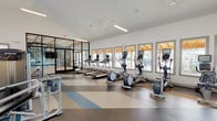This fitness center is a thoughtfully designed, single-floor facility that offers a comprehensive workout experience across several distinct zones. The space features high vaulted ceilings that contribute to an open, airy atmosphere, complemented by abundant natural light streaming through a generous row of windows along the main cardio area. The floor throughout is finished in a striking geometric pattern of blue, gray, and tan tones, adding a dynamic visual element to the space.

The cardio zone is well-appointed with multiple treadmills, elliptical machines, and stationary bikes, all positioned to face the exterior windows for an engaging workout experience. A dedicated free weights area features a full rack of dumbbells, kettlebells, and a weight bench, anchored by floor-to-ceiling mirrors that amplify the sense of space. An additional functional training zone accommodates stretching, stability work, and accessory equipment including resistance bands, stability balls, and a back extension bench.

A standout feature is the enclosed cycling studio, separated from the main floor by sleek black-framed glass partition walls and doors. This private room is highlighted by a bold teal starburst mural accent wall, a mounted TV, and a mirrored wall, creating an energizing and immersive environment for cycling classes. The overall design is clean, modern, and purposefully organized to support a wide range of fitness activities.