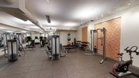 This fitness facility is designed to accommodate a comprehensive workout experience within a single-level layout. The space features rubber tile flooring throughout, providing both durability and safety for a variety of exercise activities. Overhead, a combination of track lighting and suspended linear fixtures ensures the room is well-illuminated, complementing the light-toned walls and ceiling.

The gym is outfitted with a notable selection of equipment, including a cable/functional trainer machine, adjustable weight benches, a dumbbell rack with a full range of free weights, and stability balls for core training. Cardio equipment, including a treadmill and additional machines, lines the mirrored wall along one side, which serves to visually expand the space and assist users in monitoring their form during exercise.

Exposed ductwork along the ceiling adds an industrial element to the design, while a warm wood-paneled accent wall near the entrance introduces a contrasting texture. The layout is organized to allow for comfortable movement between equipment zones, making the space functional for both individual and small-group training sessions.