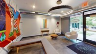 Located at 2323 NE Jackson St in Minneapolis, Minnesota, this single-floor lobby space serves as a welcoming entry point for residents. The interior showcases a contemporary industrial aesthetic, with polished concrete floors, exposed ductwork running along the ceiling, and clean white walls that create a bright, modern atmosphere.

The lounge area is a standout feature, furnished with neutral-toned armchairs, a curved sofa, and a round wooden coffee table, all anchored by a bold circular pendant light fixture that adds a dramatic focal point to the space. A colorful abstract painting adorns the wall, complementing the vibrant character of the lobby.

One of the most striking elements is a large, vivid mural celebrating the local Northeast Minneapolis neighborhood, incorporating iconic imagery such as the Grain Belt Beer sign, the Stone Arch Bridge, and a bold "NEX" lettering design in eye-catching colors. This piece grounds the space in its local identity and adds significant visual energy.

A glass-walled community room is accessible from the main lobby, offering views to the outdoors and featuring communal seating and workspace with ample natural light. Adjacent to this, a bank of resident mailboxes and elevator access provide practical functionality. The overall design balances community-oriented amenities with a gallery-like aesthetic, making for a thoughtfully curated and welcoming arrival experience.