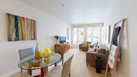 This thoughtfully designed 1-bedroom, 1-bathroom residence is laid out across a single floor, offering a comfortable and efficient use of space. The entry hallway, accented with vibrant abstract artwork, transitions seamlessly into the open living and dining area. The living room is bright and airy, featuring large windows with slatted blinds that invite abundant natural light, plush wall-to-wall carpeting, and a wall-mounted mini-split HVAC unit for year-round comfort. A defined dining area accommodates a round glass-top table, while a media console anchors the living space. The bedroom is generously proportioned, featuring soft carpet flooring, a large window, its own mini-split HVAC unit, and ample room for a full furniture arrangement. A dedicated closet with wire shelving provides practical storage. The bathroom is clean and functional, featuring a full tiled tub-shower combination with a decorative tile accent border and a curved shower rod. Throughout the residence, crisp white walls, neutral tones, and consistent carpeting create a cohesive, modern ambiance that feels both welcoming and well-organized.