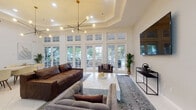 This thoughtfully designed clubhouse offers a sophisticated and welcoming environment across a single floor, combining comfort, entertainment, and refined aesthetics. The main lounge area serves as the centerpiece of the space, featuring soaring tray ceilings, a striking multi-arm chandelier, and an expansive wall of white-framed French doors and transom windows that flood the room with natural light and provide views of the outdoor pool area. Plush seating, including a rich brown leather sofa and velvet accent chairs, surrounds a substantial marble coffee table, creating an inviting gathering space. A wall-mounted flat-screen television and a sleek console table complete the lounge.

Adjacent to the main lounge, a dedicated billiards room showcases a full-sized pool table set atop a vibrant patterned rug, illuminated by a dramatic double-ring gold chandelier. A separate recreation room provides additional entertainment with a foosball table, casual seating, and decorative shelving. The dining area offers a bright and airy setting with a light wood table, cream-toned upholstered chairs, and elegant wall art, all beneath a sculptural pendant light. The entry features tall French doors with transom windows, reinforcing the open and luminous character found throughout this well-appointed clubhouse.