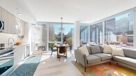 This thoughtfully designed single-floor urban residence blends contemporary architecture with an abundance of natural light. The open-concept living and dining area is anchored by expansive floor-to-ceiling windows that frame dynamic city skyline views, flooding the space with daylight throughout the day. Light hardwood flooring runs continuously through the main living areas, enhancing the bright, airy atmosphere. A sleek, modern pendant light fixture adds a stylish focal point above the dining area.

The kitchen features light wood cabinetry extending to the ceiling, white subway tile backsplash, and a full suite of stainless steel appliances, including a refrigerator, range, and microwave. Clean lines and recessed lighting give the kitchen a polished, functional aesthetic.

The primary bedroom is a calm retreat with crisp white walls, large windows offering neighborhood rooftop views, and ample room for comfortable furnishings. The en-suite bathroom is finished with a wood-toned floating vanity, white quartz countertop, undermount sink, and large mirror. A second bedroom also enjoys generous window exposure with views of the surrounding cityscape.

A second full bathroom features a combined tub and shower with tile surround and a matching wood-toned vanity. A dedicated in-unit laundry closet with a stacked washer and dryer adds everyday convenience near the entry foyer.