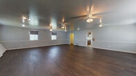This single-floor space is designed to accommodate a variety of functions, centered around a generously sized open main hall that serves as the heart of the layout. The hall features rich, dark hardwood flooring that extends throughout, providing a polished and durable surface well-suited for events, gatherings, or active use. Light gray walls are accented by a continuous white chair rail that runs the perimeter, lending a clean and structured aesthetic to the interior.

Multiple ceiling fans are evenly distributed across the ceiling, ensuring consistent airflow throughout the expansive room. Supplemental track and spotlight fixtures provide additional illumination, complementing the natural light that enters through several windows dressed with patterned valances. The space is equipped with clearly marked exit doors, indicating its suitability for public or semi-public use.

A smaller secondary room, accessible from the main hall, offers additional flexible space and mirrors the same flooring and wall finishes. Folding chairs and tables are stored within the space, highlighting its event-ready functionality.

Adjacent to the main hall, a compact kitchen features warm wood cabinetry, a gas range, microwave, refrigerator, and a double-basin sink with light countertops—providing practical support for catered events or everyday use.