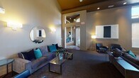 This well-appointed single-floor communal space blends contemporary design with comfort, offering multiple distinct zones for relaxation, dining, and socializing. The main lounge area features soaring ceilings with warm tray accents and recessed lighting, creating an inviting atmosphere. Plush, slate-blue sofas accented with teal and navy velvet throw pillows are arranged around glass-topped coffee tables, while a round mirror and stylish wall sconces add a refined decorative touch. A dramatic fireplace anchors one side of the room, complemented by an arched window that draws in natural light. The dining area is furnished with multiple tables and chairs beneath a striking sputnik-style chandelier, making it well-suited for gatherings. The full kitchen is equipped with rich, dark espresso cabinetry, granite countertops, a dark tile backsplash, black appliances including a refrigerator and dishwasher, dual sinks, and a breakfast bar with seating — offering full functionality for catered events or resident use. Warm tile flooring anchors the kitchen, transitioning to plush carpeting throughout the lounge and dining spaces. Throughout the space, thoughtful lighting, curated artwork, and cohesive teal and navy accents tie the design together into a polished, welcoming environment.