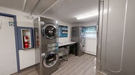 Located at 83 Mansfield Rd in New London, Connecticut, this shared laundry facility is a single-floor utility room designed for efficient, practical use. The space is outfitted with multiple stainless steel stacked washer and dryer units arranged along the walls, maximizing capacity within a compact footprint. The layout creates a clear central aisle for easy navigation and access to all machines.

The room features durable wood-look vinyl plank flooring that is easy to maintain, complemented by a textured ceiling with overhead fluorescent lighting that keeps the space well-lit. A window along one wall, dressed with a patterned valance, allows for natural light and ventilation. A built-in baseboard heating system ensures year-round comfort.

A central folding table provides a convenient surface for sorting and organizing laundry, while a rolling laundry cart adds to the room's functionality. Informational signage is posted on the walls to assist users with machine operation. The room also includes a fire extinguisher and safety notices, reflecting attention to occupant safety.

Two doors provide access points to the room. The neutral-toned walls, accented with a blue feature wall at the far end, give the space a clean and organized appearance. Overall, this laundry room is a well-maintained, fully functional shared facility designed to serve its users efficiently.