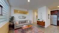 This single-floor unit showcases a cohesive mid-century modern aesthetic carried throughout its well-organized layout. The living room serves as the central gathering space, featuring terrazzo-style flooring, crisp white walls, and a distinctive glass block window wall that filters abundant natural light while maintaining privacy. A unique interior pass-through window with a yellow-accented recess connects the living area to adjacent spaces, adding architectural character. The living room opens to a dedicated dining area, which benefits from its own glass block window and includes a wall-mounted mini-split climate control unit.

The kitchen is thoughtfully designed with rich dark wood cabinetry, white countertops, a pale blue subway tile backsplash, and stainless steel appliances. A peninsula with an integrated sink and seating for two is illuminated by striking gold pendant lights, adding warmth and functionality. A full bathroom features a floating wood vanity, terrazzo flooring, and clean white finishes. The bedroom is accessible via a hallway, which also includes a quiet nook with hardwood-style flooring and a small clerestory window. Overall, the unit blends practical design with curated mid-century details for a polished, livable atmosphere.