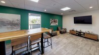 This contemporary single-floor workspace is designed to balance productivity and comfort through a cohesive blend of modern aesthetics and functional furnishings. The space is immediately distinguished by its bold teal accent walls, which contrast crisply against neutral white surfaces and light-toned carpet flooring throughout.

The main work area is equipped with a long communal desk featuring ergonomic black rolling chairs arranged alongside a striking live-edge wooden high-top table paired with industrial-style bar stools — offering versatile seating arrangements suited for both individual focus and group collaboration. Decorative hexagonal wooden shelves mounted on a white wall add a warm, organic touch to the workspace.

A lounge corner provides a relaxed seating option with upholstered armchairs, a natural wood stump side table, and a glass-topped end table, creating an inviting area for informal meetings or downtime. A wall-mounted flat-screen display is conveniently positioned nearby.

Natural light filters in through windows that offer views of surrounding greenery, enhancing the open, airy feel of the space. Recessed lighting and flush panel ceiling fixtures ensure consistent, well-distributed illumination throughout. Large-format black-and-white landscape photography displayed on acrylic mounts adds an artistic, refined element to the overall design. The space presents a clean, professional environment well-suited for modern work needs.