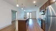 Located at 3750 Philemon Ave in Charlotte's 28205 zip code, this contemporary single-floor residence offers a thoughtfully designed open-concept layout. The kitchen serves as the centerpiece of the living area, showcasing rich dark-wood cabinetry, granite countertops, a large center island with a built-in sink, and a full suite of stainless steel appliances including a range, microwave, and refrigerator. Stylish pendant lighting illuminates the island, while light-toned hardwood flooring extends throughout the main living areas, adding warmth and visual continuity. The living space opens directly to a private balcony through glass-paned doors, offering views of the surrounding neighborhood and allowing natural light to fill the room. The bedrooms feature plush carpeting and generous walk-in closets with built-in shelving and hanging rods. Corner bedroom windows on two walls invite abundant natural light and neighborhood views, complemented by a ceiling fan for added comfort. The bathroom is well-appointed with a granite-topped vanity, hardwood-style flooring, and a glass-enclosed walk-in shower with white tile surround and brushed gold fixtures. Clean white walls and recessed lighting throughout lend the space a bright, modern aesthetic.