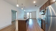 Located at 3750 Philemon Ave in Charlotte's 28205 zip code, this contemporary single-floor residence offers a thoughtfully designed open-concept layout. The kitchen serves as the centerpiece of the living area, showcasing rich dark-wood cabinetry, granite countertops, a large center island with a built-in sink, and a full suite of stainless steel appliances including a range, microwave, and refrigerator. Stylish pendant lighting illuminates the island, while light-toned hardwood flooring extends throughout the main living areas, adding warmth and visual continuity. The living space opens directly to a private balcony through glass-paned doors, offering views of the surrounding neighborhood and allowing natural light to fill the room. The bedrooms feature plush carpeting and generous walk-in closets with built-in shelving and hanging rods. Corner bedroom windows on two walls invite abundant natural light and neighborhood views, complemented by a ceiling fan for added comfort. The bathroom is well-appointed with a granite-topped vanity, hardwood-style flooring, and a glass-enclosed walk-in shower with white tile surround and brushed gold fixtures. Clean white walls and recessed lighting throughout lend the space a bright, modern aesthetic.
