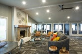 This single-story space is thoughtfully designed to serve as a shared gathering and amenity area. The layout flows openly between distinct functional zones, all unified by a cohesive modern aesthetic and a neutral gray color palette.

The kitchen area is outfitted with stainless steel appliances, gray shaker-style cabinetry, a dark slate tile floor, and a subway tile backsplash. A large center island with seating provides a casual dining surface, while a sliding glass door and a large picture window offer direct views and access to an outdoor pool area. A dedicated dining table with metal bistro-style chairs accommodates group meals nearby.

Transitioning into the lounge, the space opens up dramatically beneath vaulted, beam-accented ceilings. A ceiling fan and recessed lighting illuminate the room, while multiple windows fill it with natural light and pool views. A statement stone fireplace with a natural wood mantel serves as the focal point, flanked by leather accent chairs and a substantial sofa. The patterned, wood-look flooring adds texture and warmth throughout.

A glass-windowed private office is accessible from the lounge, providing a quieter workspace within the broader communal setting. Overall, the space strikes a balance between functional utility and inviting, contemporary design.