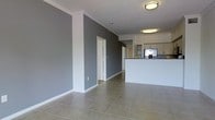This single-floor space presents a clean, modern aesthetic defined by a neutral grey and white color palette throughout. The open-plan living and kitchen area creates a sense of spaciousness, with large ceramic tile flooring laid in a grid pattern extending across the entire space. The walls are finished in a bold charcoal grey, accented by crisp white crown molding and baseboards that provide a sharp contrast and architectural definition to each room.

The kitchen is positioned at one end of the open-plan area and features a raised breakfast bar counter finished in white, providing a natural visual divider between the kitchen and living zones. White cabinetry lines the upper kitchen walls, offering ample storage, while a stainless steel refrigerator adds a contemporary touch. Flush-mounted ceiling light fixtures provide consistent overhead illumination throughout the space.

A doorway visible from the main living area leads to additional rooms, suggesting a functional layout beyond the open-plan zone. The overall ambiance is modern and minimalist, with the consistent use of grey tones, white trim, and tile flooring creating a cohesive and easily maintainable interior. The space is well-suited for those who appreciate a contemporary, low-maintenance design with an open and connected feel.