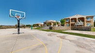 This expansive outdoor amenity area serves as a central recreational hub for its surrounding multi-story residential community, which features warm stucco exteriors, columned entryways, and stone retaining walls that lend the complex a classic Southwestern character. The grounds are thoughtfully designed to offer a variety of activities and gathering spaces beneath open desert skies.

At the heart of the space is a full-size concrete basketball court, clearly marked with painted lines and equipped with regulation backboards. Adjacent to it, a dedicated volleyball court with a net post provides additional athletic options. Both courts are oriented to take advantage of the open surroundings, with unobstructed views of the surrounding high-desert landscape and distant mountain ranges.

Expansive, well-maintained green lawns stretch between the courts and the residential buildings, offering open space for relaxation or informal recreation. A covered columned pavilion with staircase access connects the grounds to the clubhouse, while a shaded pergola structure at the far end of the property provides a picnic and seating area with desert scenery as a backdrop. Benches, trash receptacles, and mature trees are thoughtfully placed throughout, enhancing the functionality and comfort of this vibrant outdoor community space.