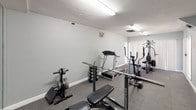 This dedicated fitness room offers a fully functional workout environment designed to support a comprehensive exercise routine. The space is finished with soft sage-green walls and durable rubber interlocking floor tiles, providing both a practical and visually cohesive atmosphere suited for regular training use.

The room is well-lit by multiple fluorescent strip lights mounted on the textured ceiling, ensuring consistent, even illumination throughout. Ventilation is provided via ceiling and wall-mounted HVAC vents, maintaining comfortable air circulation during workouts.

Equipment in the space includes a multi-station cable weight machine, a treadmill, an elliptical trainer, a stationary cycle, a adjustable weight bench with barbell and rack, a dumbbell rack with free weights, and additional resistance and cardio accessories. A large wall mirror on one side enhances the sense of space while allowing users to monitor form during exercise.

Two white paneled doors provide access points, and white baseboards frame the perimeter cleanly. Wall-mounted accessories, including a glove or accessory holder and a hand sanitizer dispenser, add functional convenience. Overall, the room is organized for efficient use, offering a self-contained and well-appointed private gym setup.