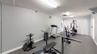 This dedicated fitness room offers a fully functional workout environment designed to support a comprehensive exercise routine. The space is finished with soft sage-green walls and durable rubber interlocking floor tiles, providing both a practical and visually cohesive atmosphere suited for regular training use.

The room is well-lit by multiple fluorescent strip lights mounted on the textured ceiling, ensuring consistent, even illumination throughout. Ventilation is provided via ceiling and wall-mounted HVAC vents, maintaining comfortable air circulation during workouts.

Equipment in the space includes a multi-station cable weight machine, a treadmill, an elliptical trainer, a stationary cycle, a adjustable weight bench with barbell and rack, a dumbbell rack with free weights, and additional resistance and cardio accessories. A large wall mirror on one side enhances the sense of space while allowing users to monitor form during exercise.

Two white paneled doors provide access points, and white baseboards frame the perimeter cleanly. Wall-mounted accessories, including a glove or accessory holder and a hand sanitizer dispenser, add functional convenience. Overall, the room is organized for efficient use, offering a self-contained and well-appointed private gym setup.