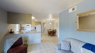 This single-story space features an open-concept design that seamlessly connects the kitchen, dining area, and living room into one cohesive living environment. Light-toned ceramic tile flooring runs throughout, lending a clean, consistent look across the entire space.

The kitchen is positioned along one side and is outfitted with light wood cabinetry, white appliances including a refrigerator, microwave, and range, as well as a countertop peninsula that provides additional prep space and serves as a natural divider between the kitchen and the adjacent living areas. A single-basin sink sits beneath overhead cabinetry, maximizing storage.

The dining area is situated just beyond the kitchen and is anchored by a pendant light fixture hanging above a wooden dining table surrounded by matching chairs, creating a warm and functional space for meals. A built-in shelving niche in the adjacent wall adds both character and display storage.

The living area is defined by soft blue accent walls that contrast with the neutral tones found elsewhere in the space. A textured ceiling adds subtle visual interest overhead. Overall, the layout feels open and efficiently organized, with a calm, understated ambiance throughout.