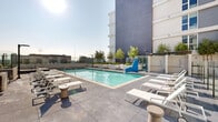 This outdoor amenity level is set within a striking contemporary multi-story residential complex, characterized by a clean, modern architectural palette of white stucco, dark gray paneling, and large-format glazing with private balconies overlooking the shared spaces below. The centerpiece is a generously proportioned swimming pool with vivid turquoise water, tiled edges, and an adjacent spa, all surrounded by a wide pool deck finished in large-format concrete pavers. Rows of white chaise lounges line the pool deck, providing ample space for sunbathing, while glass perimeter railings offer open city views beyond. A separate courtyard area features an open-sky design framed by the surrounding building wings, furnished with woven lounge chairs and built-in concrete bench seating arranged around landscaped planters. Slender ornamental trees are distributed throughout, adding a natural, softening element to the hardscape. An outdoor dining area provides wooden communal tables and chairs alongside a built-in bar counter, well-suited for social gatherings. Curved planter walls with ground-level accent lighting add architectural interest throughout the courtyards. The overall ambiance is bright, airy, and resort-inspired, blending urban sophistication with comfortable outdoor living.