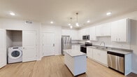 This well-appointed single-floor residence showcases a thoughtfully designed open-concept layout with a clean, contemporary aesthetic throughout. The kitchen serves as a standout centerpiece, featuring crisp white shaker cabinets, light granite countertops, and a full suite of stainless steel appliances including a refrigerator, electric range, microwave, and dishwasher. A central island with matching granite provides additional prep space and casual seating, while elegant pendant lighting adds a refined touch overhead. Light-toned hardwood-style flooring flows seamlessly through the kitchen and living areas, enhancing the sense of openness and continuity. The living area is bright and welcoming, benefiting from natural light through a large window and a glass-panel door that opens to an outdoor space, offering pleasant exterior views. The bedroom is finished with plush wall-to-wall carpeting and features a generously sized window that fills the room with natural light. The bathroom is well-equipped with a dual-sink vanity topped with granite countertops, a large mirror, and a glass-enclosed shower. Neutral wall tones and consistent finishes throughout the space create a cohesive, move-in-ready environment that balances comfort with modern style.