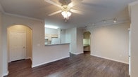 This single-story residence in Duncanville offers an open and functional layout designed for comfortable everyday living. The main living area features rich, dark hardwood-style flooring that extends throughout, providing a warm and cohesive aesthetic. Crown molding lines the ceiling, adding a refined architectural detail, while a ceiling fan with a light fixture serves as a practical central fixture in the space.

The kitchen is partially open to the living area via a raised breakfast bar, offering a natural divide without fully closing off the space. Light-toned cabinetry, a white refrigerator, and under-cabinet fluorescent lighting are visible, providing a clean and functional cooking environment. The dark countertop of the bar offers a subtle contrast to the kitchen's lighter elements.

A distinctive arched doorway leads from the main living area into a hallway, suggesting access to additional rooms within the unit. Track lighting is mounted near this transitional area, adding supplementary illumination. The overall color palette is neutral, with soft beige walls throughout, creating a bright and versatile environment that adapts well to a variety of interior styles.