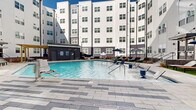 Located at 120 Matheson Ave in Charlotte, NC, this outdoor amenity space showcases a generously sized swimming pool nestled within a courtyard formed by a contemporary multi-story residential building. The pool features clean, crystal-clear water and is surrounded by a broad, light-toned stone tile deck that provides ample space for lounging and movement. The sundeck is well-appointed with a collection of lounge chairs — including distinctive orange and navy options — along with shade umbrellas in both solid navy and striped black-and-white patterns, offering a comfortable outdoor retreat in varying weather conditions. A wooden pergola structure adds an architectural accent to one side of the pool area, while raised planters with ornamental grasses and decorative rock landscaping provide natural texture and definition along the perimeter. A utility building with a secure entrance is integrated into the courtyard design. The surrounding building exterior features a modern aesthetic with a combination of white and dark gray siding, large uniformly spaced windows, and brick accents. The open-sky courtyard orientation allows for abundant natural light throughout the day, creating a bright and inviting atmosphere ideal for outdoor recreation and relaxation.