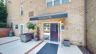 Radwyn's amenity level offers a thoughtfully designed, single-floor space that seamlessly blends community, productivity, and wellness. The entrance, framed by a classic brick exterior and a clean canvas awning bearing the Radwyn name, welcomes residents with planters and a covered walkway leading into a bright, modern lobby. Large-format tile flooring, recessed lighting, and glass doors opening to an outdoor patio create an inviting, open feel throughout.

The lounge area serves as the social heart of the space, distinguished by a sweeping, large-scale abstract mural in warm earth tones — yellows, oranges, and greens — that enlivens the entire room. Comfortable seating, a bold orange area rug, and a sculptural coffee table complete the setting, while a wall-mounted display and a coffee station add practical convenience.

Adjacent to the lounge, glass-walled conference rooms provide transparent, professional meeting spaces outfitted with wooden tables, ergonomic chairs, and pendant lighting — ideal for focused work or collaborative sessions.

The expansive fitness center rounds out the amenities, featuring rubber flooring, floor-to-ceiling mirrors, rows of cardio machines including treadmills and ellipticals, a full complement of strength-training equipment, and free weights — all arranged to maximize usability within a generously sized, well-lit environment.