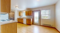 This single-floor space offers a functional and well-organized layout designed for comfortable everyday living. The kitchen is a standout feature, equipped with warm honey-toned oak cabinetry, white appliances including a range, refrigerator, microwave, and dishwasher, all set atop light hardwood flooring that extends seamlessly into the open living area. A double sink sits beneath a window, and a glass-paneled exterior door fills the kitchen with natural light while providing direct outdoor access. The open-concept living and dining area is bright and spacious, with clean white walls and the same light hardwood floors creating a cohesive, airy feel. A dedicated laundry room is conveniently located just off the kitchen, housing a full-sized washer and dryer with wire shelving above for additional storage. The bedrooms feature neutral carpet flooring and blinds-covered windows that let in ample natural light and offer views of the surrounding greenery. Baseboard heating is present throughout. The bathroom is equipped with accessibility features including grab bars near the toilet and shower, tile flooring, and a window for natural ventilation. Overall, the space maintains a clean, practical aesthetic with consistent finishes throughout.
