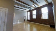 This single-floor space showcases a compelling blend of historic industrial character and modern functionality. The most striking feature is the original exposed brick wall, which spans a significant portion of the interior and is punctuated by multiple arched and rectangular windows that flood the space with natural light. The soaring ceilings draw the eye upward to reveal a combination of rustic wood beams and exposed ductwork, lending the space an authentic loft aesthetic. Polished hardwood floors run throughout, providing a warm contrast to the raw industrial elements overhead. A ceiling fan is mounted centrally, contributing to both air circulation and the overall utilitarian charm. The layout is open and expansive, offering excellent flexibility for a variety of uses. Pendant lighting is strategically placed to supplement the abundant natural light. A secondary room or enclosed area is accessible through a doorway visible in the background, adding a degree of functional versatility to the otherwise open floor plan. The overall ambiance is one of creative, adaptable space rooted in industrial heritage.