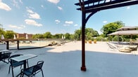 This outdoor amenity space is designed to deliver a resort-like experience, centered around a generously sized swimming pool enclosed by sleek glass railings that preserve unobstructed views of the lush, tree-lined surroundings. The expansive concrete deck provides ample room for lounging and socializing, furnished with a mix of stylish seating options including wire-frame lounge chairs, pod-style accent chairs, and bistro table-and-chair sets ideal for casual outdoor dining.

A striking dark-wood pergola anchors the space architecturally, offering partial shade and adding a refined structural element to the open-air environment. Beneath its canopy sits a fully equipped outdoor kitchen, featuring a built-in stainless steel grill set into a stacked-stone base with a granite countertop — perfect for entertaining. Additional shaded areas are provided by freestanding market umbrellas and a gazebo positioned toward the perimeter of the deck.

The adjacent building features warm brick masonry construction with large floor-to-ceiling glass doors that open directly onto the deck, connecting interior amenity spaces — including what appears to be a fitness center — seamlessly to the outdoors. An upper-level balcony overlooks the entire space, adding a sense of vertical dimension. Under sunny skies with sweeping green canopy views, this outdoor space creates an inviting and vibrant atmosphere year-round.