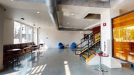 Located at 5 E Reed St in the heart of San Jose, California, this contemporary single-floor space combines industrial design with warm, inviting interiors. The expansive open-plan layout is anchored by polished concrete floors that reflect abundant natural light streaming in through large picture windows, offering views of the surrounding streetscape.

The main lounge area is a standout feature, showcasing bold yellow accent walls paired with rich wood paneling that creates a striking two-tone aesthetic. Comfortable grey sectional sofas and modern lounge chairs are arranged around a central coffee table, complemented by curated art pieces including a sculptural city-map wall installation. Exposed industrial-style ductwork running across the ceilings reinforces the space's urban, loft-inspired character.

An open staircase with metal railings and warm wood treads provides access to an upper level, serving as a visual focal point within the main floor. Adjacent to the lounge, a casual seating area with bar-height tables and chairs offers additional gathering space bathed in natural light. A warm amber-toned glass partition adds a distinctive architectural element near the staircase. The overall ambiance balances industrial architecture with curated, colorful furnishings, creating a dynamic and welcoming communal environment.
