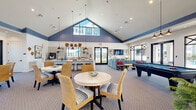 This single-floor community clubhouse offers a generously sized, open-concept layout designed for social gatherings and relaxation. The space is defined by its dramatic vaulted ceilings accented with bold navy blue trim, which contrast beautifully against crisp white shiplap walls and light hardwood flooring in the lounge area. A striking floor-to-ceiling triangular window floods the main lounge with natural light and provides views of the surrounding outdoor amenities.

The lounge area features plush navy leather swivel chairs and a comfortable sectional sofa arranged around a gold-accented coffee table, creating an inviting focal point. A large-format wall-mounted TV and curated artwork add a polished, residential feel. A sliding barn door connects the lounge to the adjacent gathering room, adding a charming architectural detail.

The adjoining social room transitions to carpeted flooring and includes casual dining tables with wicker and striped upholstered chairs, a long granite-topped bar with barstool seating, and a full pool table for recreation. A wet bar area with countertop appliances and open shelving supports entertaining needs. Large black-framed windows and glass doors line the perimeter, offering continuous outdoor views and allowing natural light to permeate the entire space.