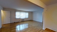 This single-floor space offers a clean and open layout highlighted by beautifully polished hardwood floors that reflect natural light throughout the interior. The main living area is spacious and airy, with smooth, neutral-toned walls that create a bright and welcoming ambiance. A large window dressed with vertical blinds serves as the focal point of the room, flooding the space with abundant natural light while offering a view of the greenery outside. A wall-mounted air conditioning unit sits beneath the window, complementing the baseboard heating that runs along the perimeter of the room — ensuring year-round climate comfort. The open-plan design allows for flexible furniture arrangement, making the living area highly functional and adaptable to a variety of uses. Architectural details include a subtle ceiling soffit that adds visual structure and depth to the space. White baseboards and clean wall finishes throughout contribute to the overall sense of freshness and simplicity. The seamless flow between areas makes the space feel cohesive and well-connected. Overall, this residence combines practical features with a light-filled, understated aesthetic that provides a comfortable and versatile living environment.