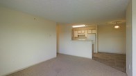 This single-floor residence offers a straightforward and functional layout designed for comfortable everyday living. The living area is finished with wall-to-wall carpet in a neutral tone, providing a clean and cohesive look that complements the light-colored walls throughout. The space feels open and uncluttered, offering a blank canvas well-suited for a variety of furnishing arrangements.

A defining feature of the layout is the pass-through kitchen, which connects to the living area via a raised counter that doubles as a breakfast bar or serving surface. The kitchen is equipped with light wood cabinetry providing ample storage, a white refrigerator, a range, and an overhead fluorescent light fixture that keeps the cooking area well-lit. The kitchen flooring transitions to tile, offering a practical, easy-to-clean surface in this high-use zone.

The textured ceiling adds a subtle architectural detail throughout, while the neutral color palette of cream and beige tones creates a warm, inviting ambiance. The overall design emphasizes practicality and livability, making efficient use of the available space. The open sightline between the living and kitchen areas enhances the sense of space and promotes a connected, sociable atmosphere within the home.