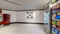 This 210-square-foot single-floor common area serves as a lobby or elevator foyer, functioning as a transitional space within a larger multi-floor building. The space is defined by large-format white ceramic tile flooring, which lends a clean, professional aesthetic throughout. Dark, contrasting baseboards and crown molding trim the walls and ceiling perimeter, creating a sharp, structured visual boundary that adds a modern, polished character to the room.

A standout architectural feature is the curved wall adjacent to the elevator bank, which introduces an elegant, dynamic form into what might otherwise be a purely utilitarian corridor. Stainless steel elevator doors are positioned along one wall, reinforcing the commercial, upscale nature of the space.

Recessed ceiling lighting provides consistent, even illumination across the room, complemented by track lighting in select areas. One wall displays a curated 3x3 grid of framed monochromatic artworks, adding a gallery-like quality and visual interest to the otherwise minimalist space.

A convenience retail kiosk — featuring glass-front refrigerators stocked with beverages and snacks alongside open shelving with packaged goods — is incorporated into the space, offering practical amenity value for building occupants. The hallway visible beyond connects to additional building corridors, confirming this area's role as a central transit and amenity hub.