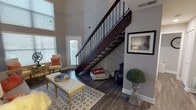 This two-story home impresses from the moment you enter the main living area, where soaring double-height ceilings create a dramatic sense of openness. A striking staircase with a dark wood finish and sleek metal balusters serves as an architectural centerpiece, anchoring the space while drawing the eye upward. Large windows flood the living room with natural light, complementing the rich, dark hardwood flooring that runs throughout the main level. The living room is generously proportioned, offering ample space for seating arrangements beneath the grand ceiling height.

The upper level opens to a versatile loft area that functions as a flexible workspace or relaxation nook, complete with its own seating and overlooking the staircase landing. Multiple bedrooms are found on this floor, including a well-appointed primary bedroom with warm, neutral tones and a secondary bedroom with carpeted flooring. Two full bathrooms feature warm wood cabinetry, white countertops, tiled tub-shower combinations accented with mosaic tile borders, and wood-look flooring. A dedicated wet bar area adds convenience, outfitted with dark granite countertops, warm wood cabinetry, a stainless steel sink, and a decorative mosaic tile backsplash — an ideal spot for entertaining.