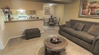 This single-floor space offers an open-concept layout that seamlessly integrates the living, dining, and kitchen areas into one cohesive and functional environment. The living area features plush carpeting and is anchored by a comfortable sofa with decorative accent pillows, an upholstered ottoman, and a round glass-top coffee table — all finished in warm, earthy tones that contribute to the space's inviting ambiance. A breakfast bar separates the kitchen from the rest of the open floor plan, providing a natural transition point while maintaining the open sightlines throughout. The kitchen features light wood cabinetry and offers counter seating along the bar. Adjacent to the kitchen, a dedicated dining area accommodates a full dining table and chairs, making it ideal for everyday meals or entertaining guests. Warm neutral wall tones and soft ambient lighting enhance the cozy atmosphere throughout the space. The consistent carpeted flooring across the main living and dining areas ties the open layout together, creating a unified and welcoming feel from every vantage point.