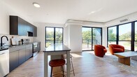 This thoughtfully designed single-floor unit showcases a clean, modern aesthetic throughout. The open-concept layout connects the kitchen and living area seamlessly, creating a spacious and functional central hub. The kitchen is outfitted with matte black cabinetry, stainless steel appliances including a refrigerator and range, a dark countertop, and a striking geometric-patterned tile backsplash. A freestanding island with bar seating provides additional prep space and casual dining. The living area is bright and inviting, anchored by large black-framed windows and a sliding glass door that opens to an outdoor balcony, framing views of mature trees and greenery. Light wood-tone flooring runs throughout the main living spaces, complementing the crisp white walls. Two bedrooms offer a comfortable retreat, each featuring plush carpeting and generous windows that bring in natural light and outdoor views. The full bathroom is finished with matte black fixtures, dark vanity cabinetry, a white countertop, and subway tile surrounding the tub and shower. Exposed ductwork adds an understated industrial touch to the living area ceiling, while the overall design balances contemporary minimalism with warm, livable details.