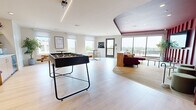 This single-floor amenity lounge offers a thoughtfully designed communal space that balances recreation, relaxation, and socializing. Light hardwood-style flooring runs throughout the main open area, which is bathed in natural light from multiple large windows and sliding glass doors that lead to an outdoor balcony, offering open exterior views.

The space is divided into distinct functional zones. A game area anchors one section, featuring a foosball table as a central recreational element. Adjacent to it, a compact kitchenette is outfitted with light wood cabinetry, a stainless steel under-counter refrigerator, and floating shelves set against a striking watercolor-style mural wall in soft aqua tones, adding an artistic focal point to the space.

A high-top bistro table with wire-frame stools provides a casual seating option near the balcony door, ideal for informal gathering. A built-in bench with storage and cushioned seating runs along the kitchenette wall, offering additional lounge space.

The most dramatic zone is a sunken media alcove defined by deep crimson vertical-panel walls and a matching ceiling, creating an intimate, theater-like atmosphere. A wall-mounted flat-screen TV, plush sectional sofa, and soft carpeting complete this cozy retreat within the larger space.